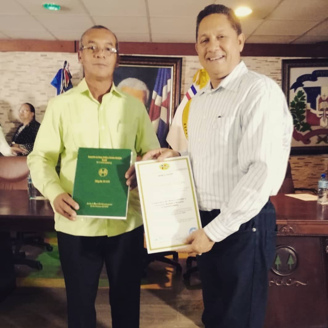 Dos hombres sonríen mientras sostienen un certificado y un libro con el símbolo cooperativo en un acto institucional.